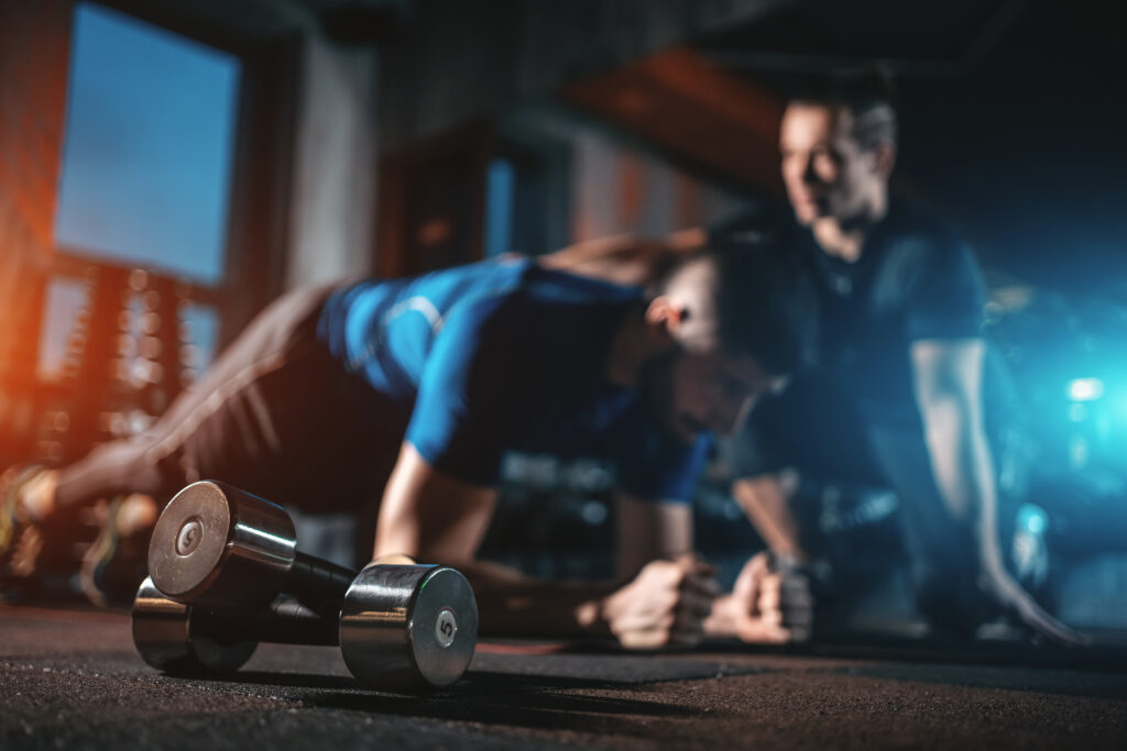 Deux hommes en plein entraînement de gainage (planche) dans une salle de sport sombre, haltères de 5 kg au premier plan.